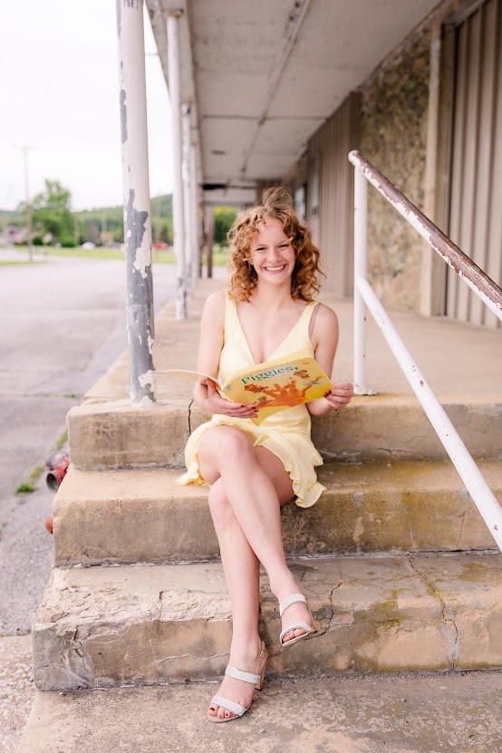 young girl with curly blonde hair in a yellow dress sitting on steps, reading a children's book 