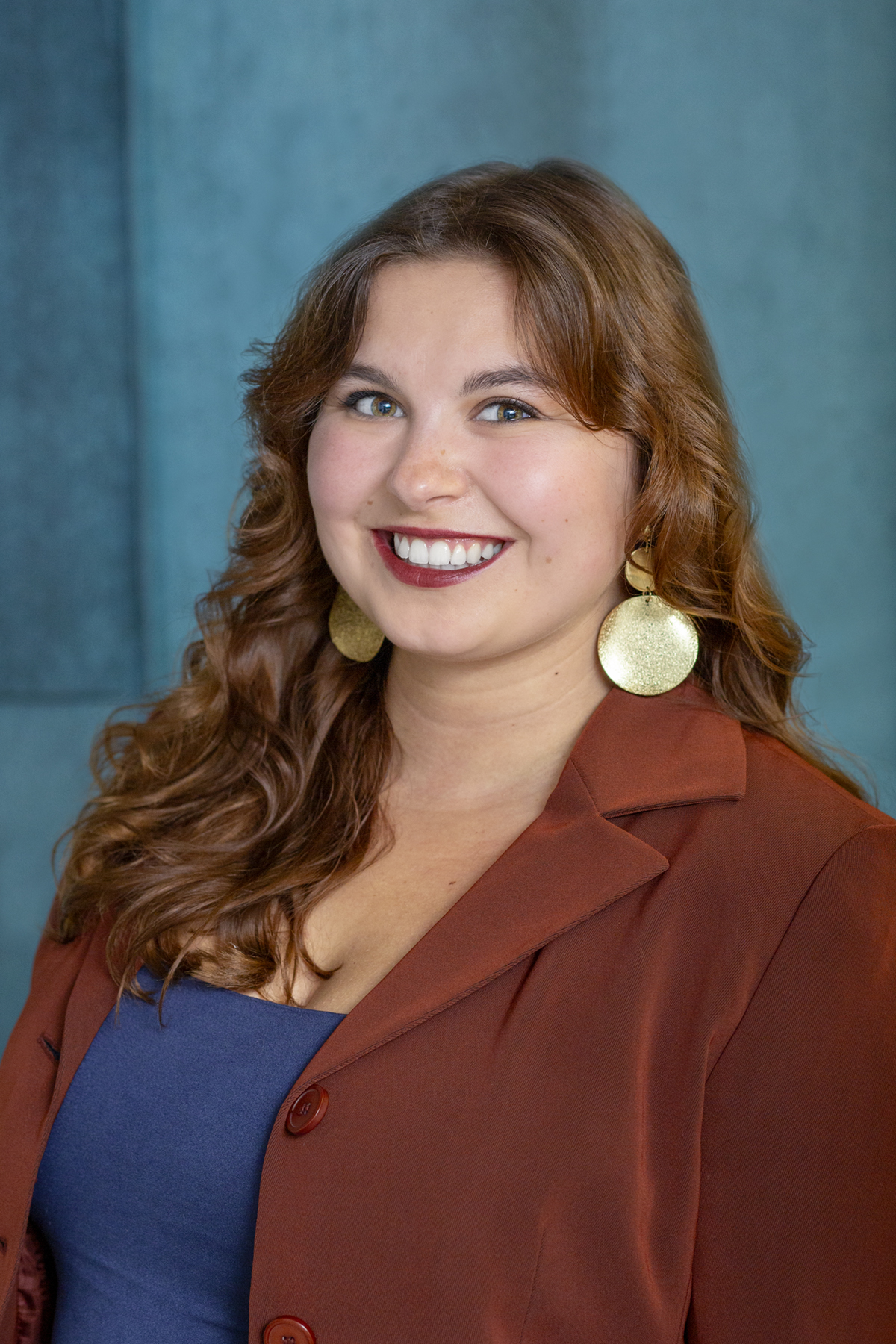 Headshot of young lady with brown hair in a brown jacket and blue shirt