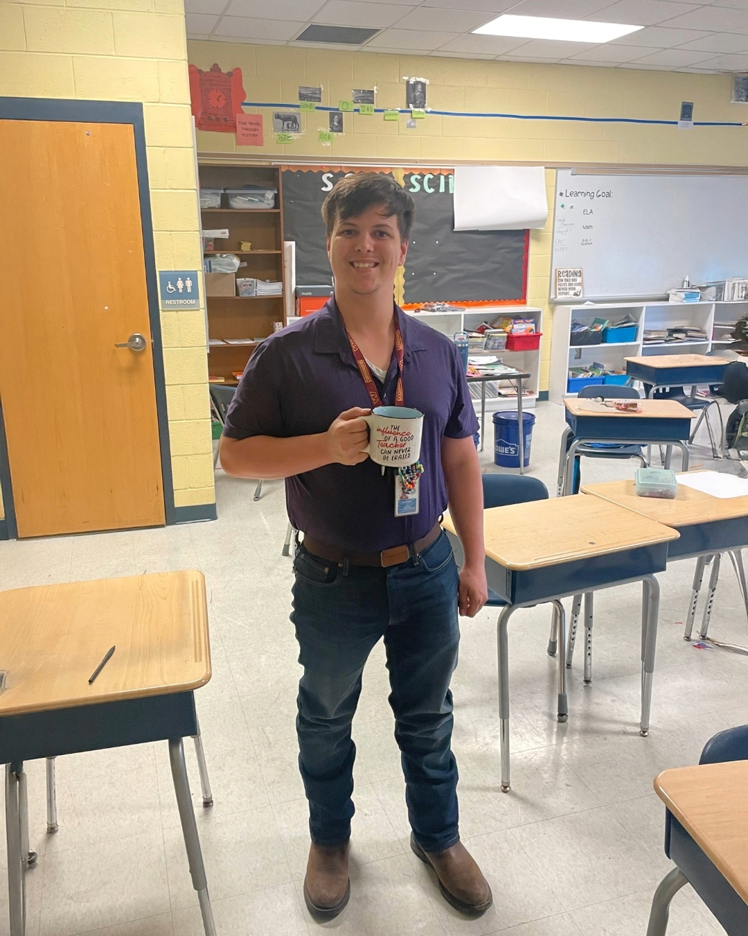 Young male teacher standing in a classroom holding a coffee mug