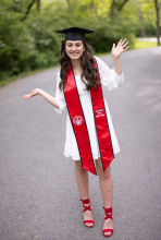 young girl with dark hair in a graduation cap, white dress and red stole.